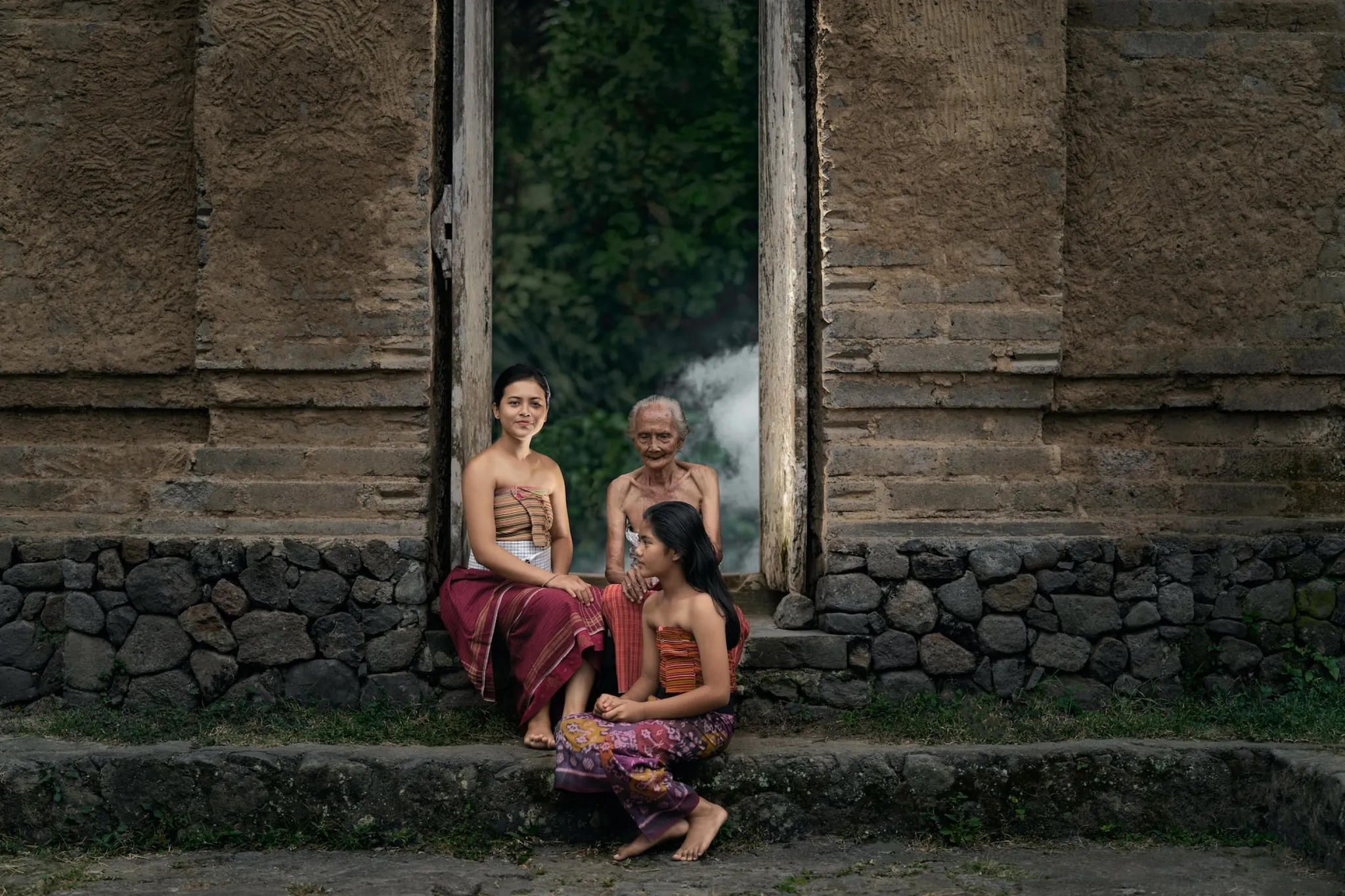 Three generations of women in traditional dress at a stone gate in Tenganan Village